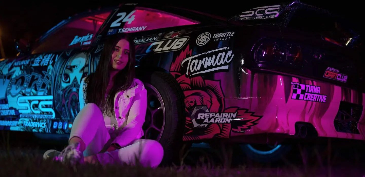 Aussie designer Tiana Semrany sitting in front of her custom-liveried drift car with a vibrant, illuminated design, showcasing her passion for motorsport and creative branding.