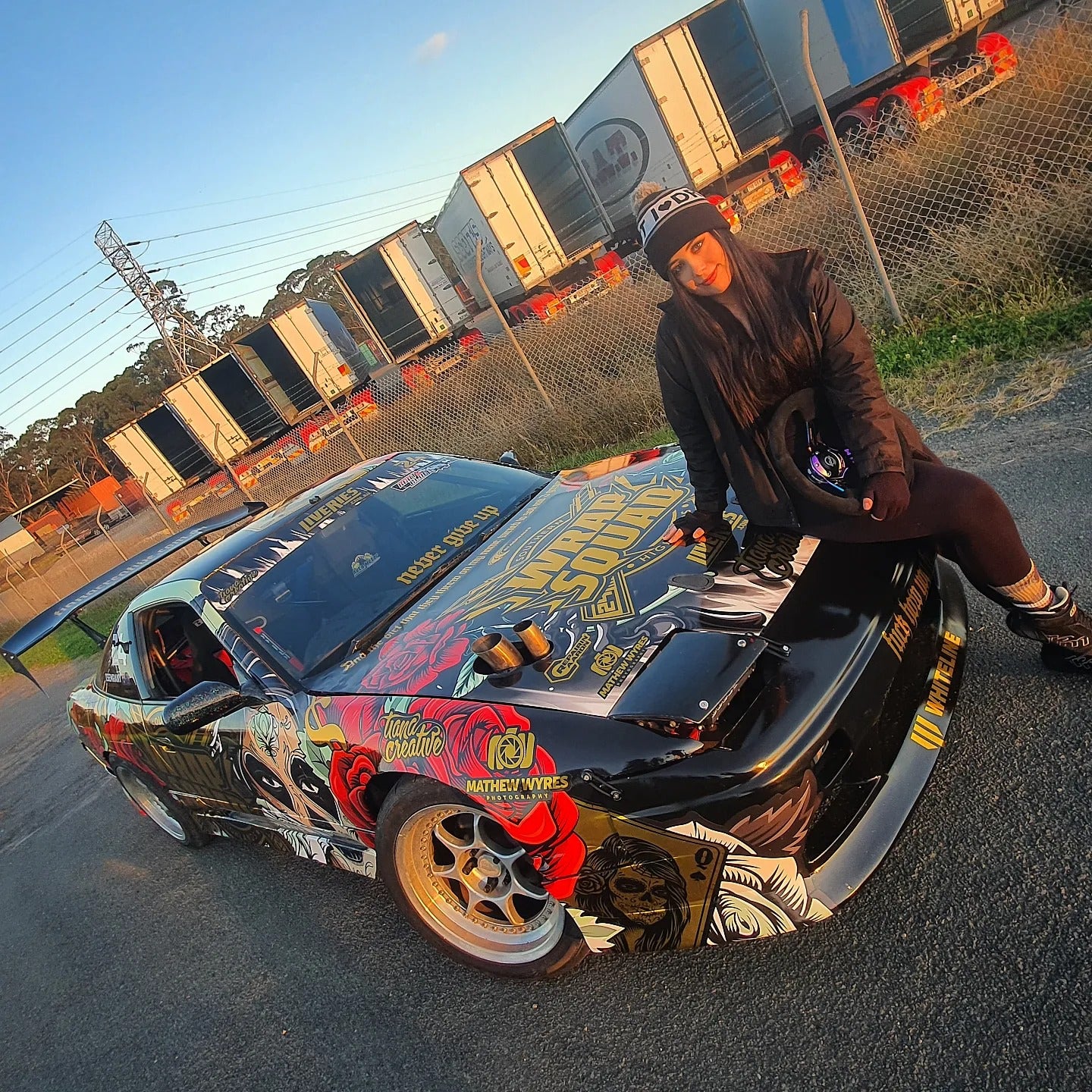 Tiana Kaiserfeld standing beside her intricately designed drift car, featuring a custom livery with vibrant artwork and branding. She holds a helmet and wears a beanie and jacket, reflecting her active involvement in the motorsport scene.