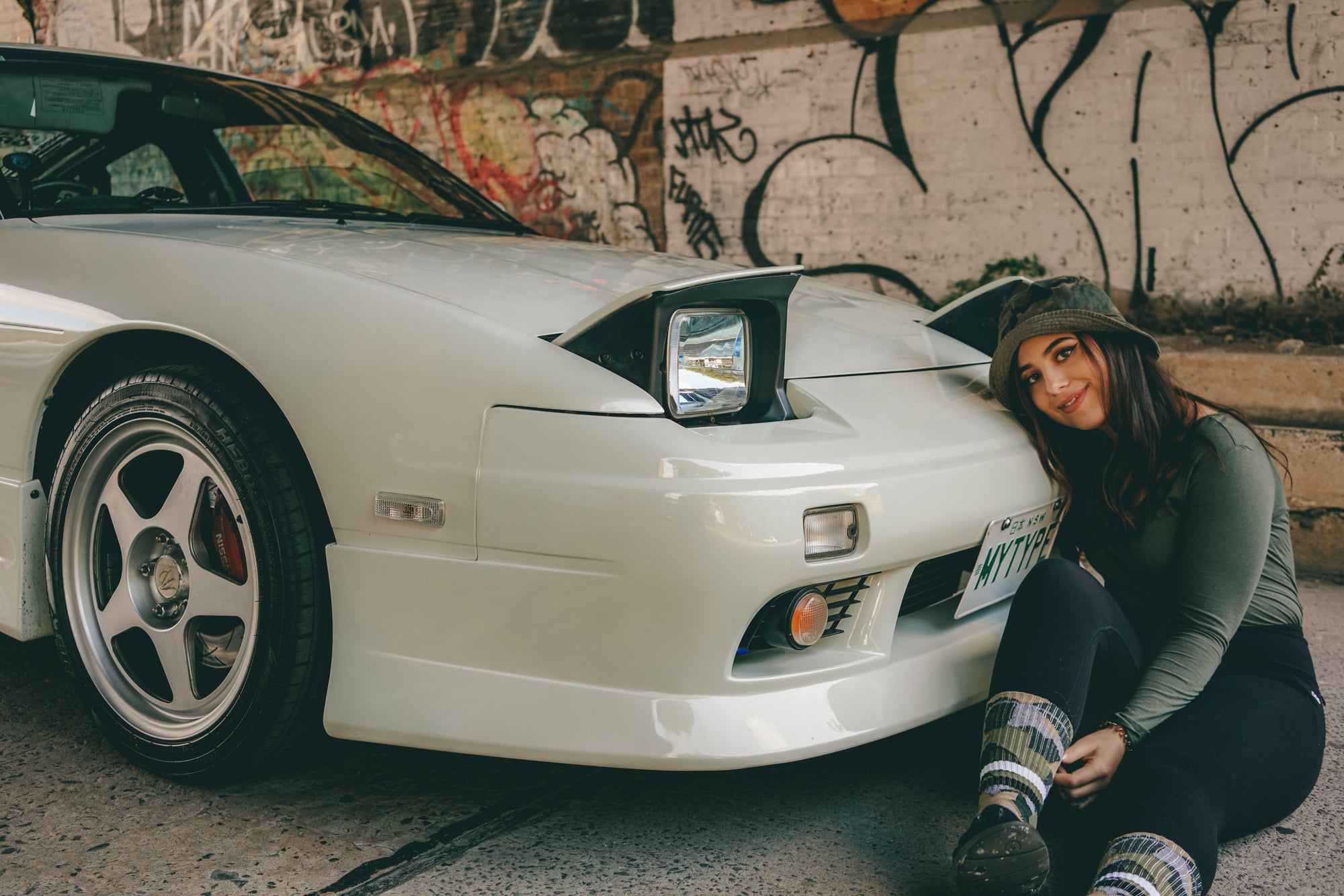 Portrait of Tiana Kaiserfeld leaning against a white car with a custom 'MYTYPE' license plate, showcasing her connection to car culture and automotive design. The setting features graffiti art in the background, reflecting a blend of urban style and personal passion for cars.