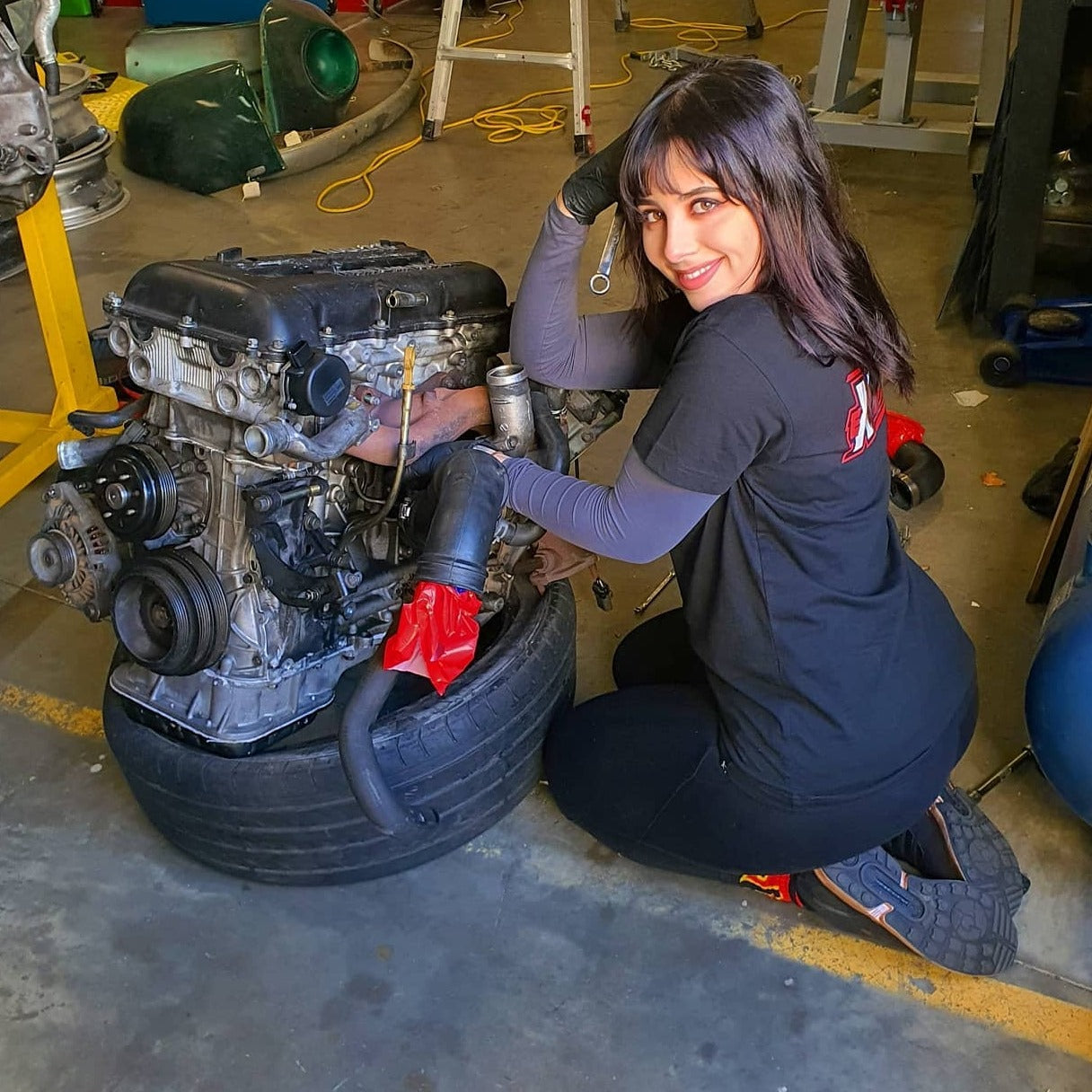 Tiana Kaiserfeld working on a car engine in a garage setting, wearing mechanic gloves and holding a wrench, showcasing her hands-on involvement in automotive mechanics. She is smiling at the camera, embodying her passion for car culture and engineering.
